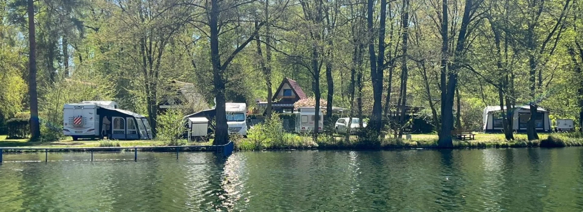Blick vom Wasser auf den Campingplatz am Steinbruchsee S&uuml;plingen mit Wohnwagen, Zelten und B&auml;umen in ruhiger, gr&uuml;ner Umgebung.