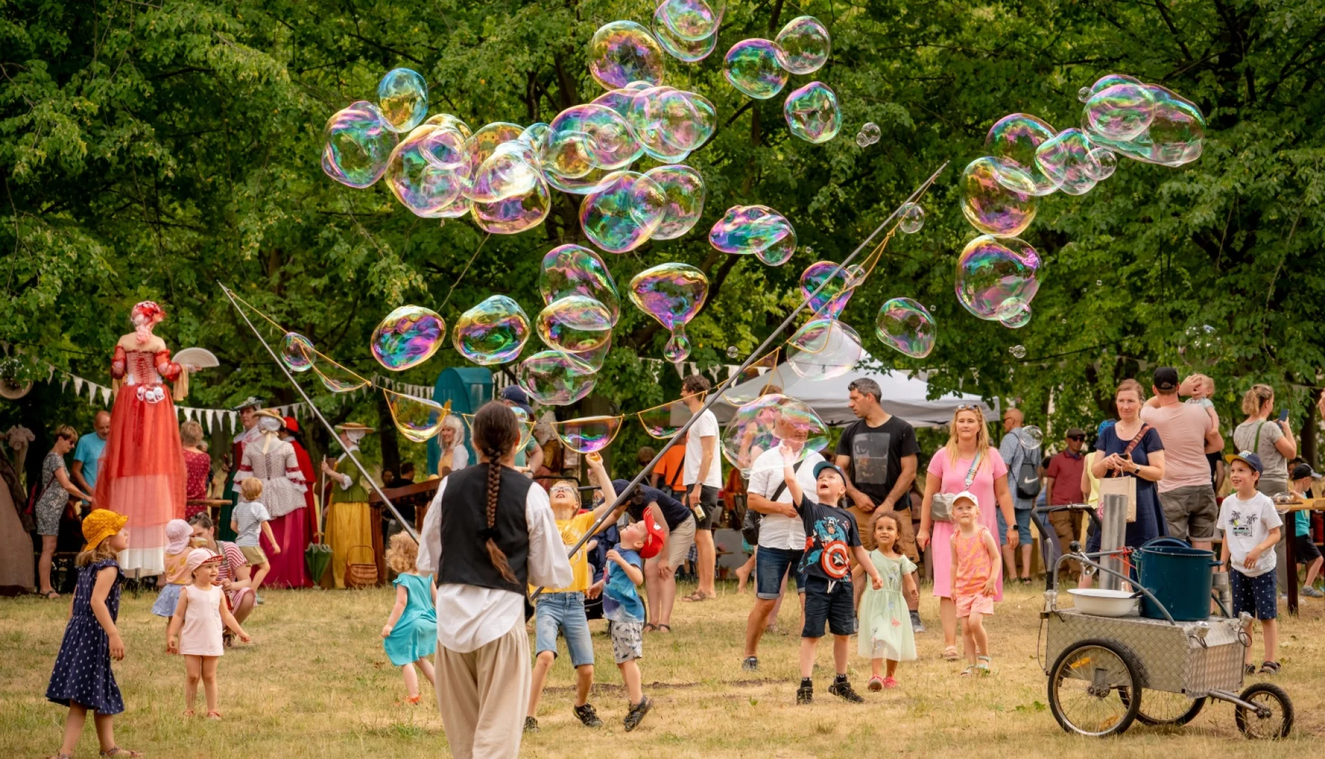 Seifenblasen-Kuenstlerin laesst dutzende schillernde Riesenblasen ueber staunende Kinder im Park schweben, dahinter Stelzenlaeuferin in Barockkostuem