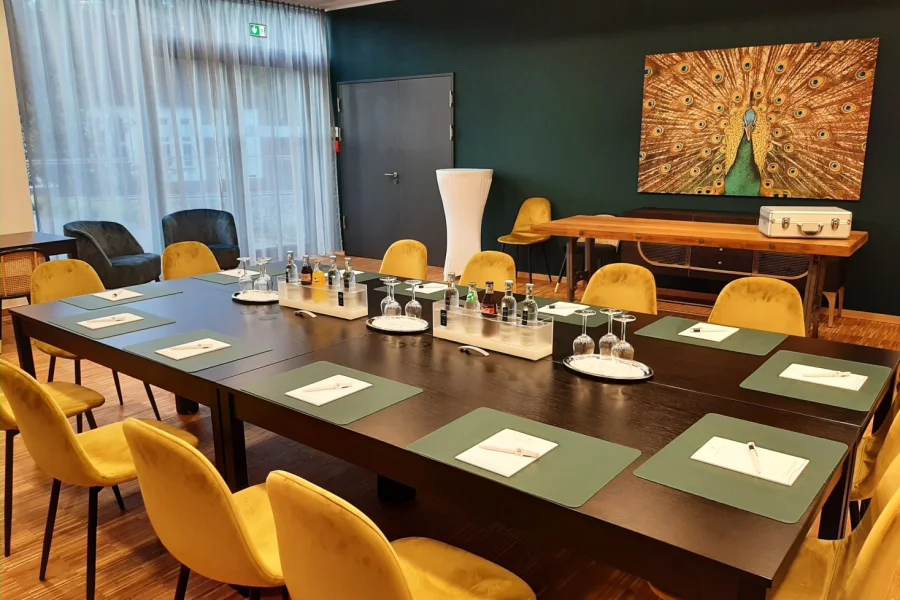 Konferenztisch aus dunklem Holz mit gr&uuml;nen Schreibunterlagen und gelben St&uuml;hlen, an der dunkelgr&uuml;nen Wand ein gro&szlig;formatiges Pfauenbild