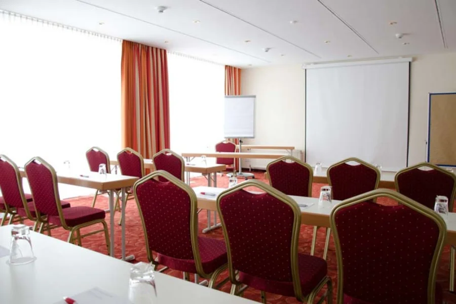 Seminarraum mit weinroten Polsterst&uuml;hlen an hellen Tischen, an der Stirnseite Leinwand und Flipchart, orange Vorh&auml;nge am Fenster