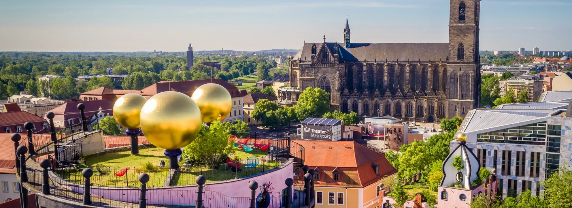 Blick ueber die Dachterrasse der Gruenen Zitadelle mit ihren goldenen Kugeln zum Magdeburger Dom unter blauem Himmel