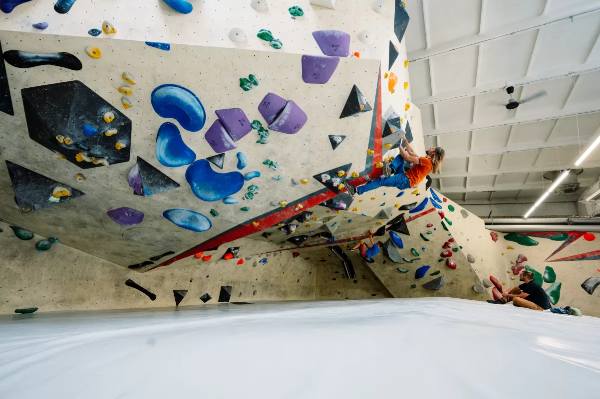 Kletterwand in der Blocschmiede Magdeburg, an der ein Mann eine Route klettert. Ein weiterer Mann sitzt auf der Matte darunter und guckt zu.