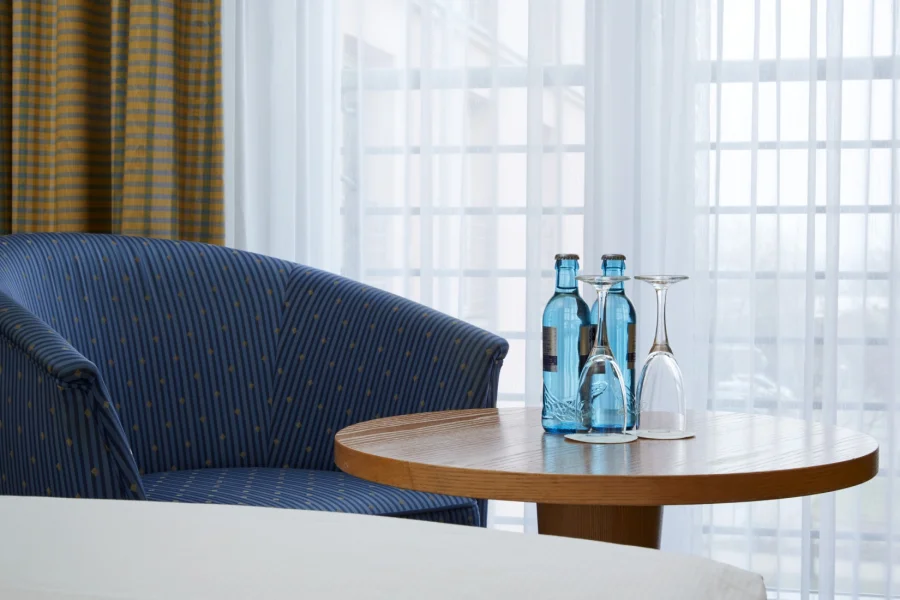 Sitzecke im Hotelzimmer: blau gestreifter Sessel neben einem runden Holztisch mit zwei Wasserflaschen und Gl&auml;sern vor einem hellen Vorhang