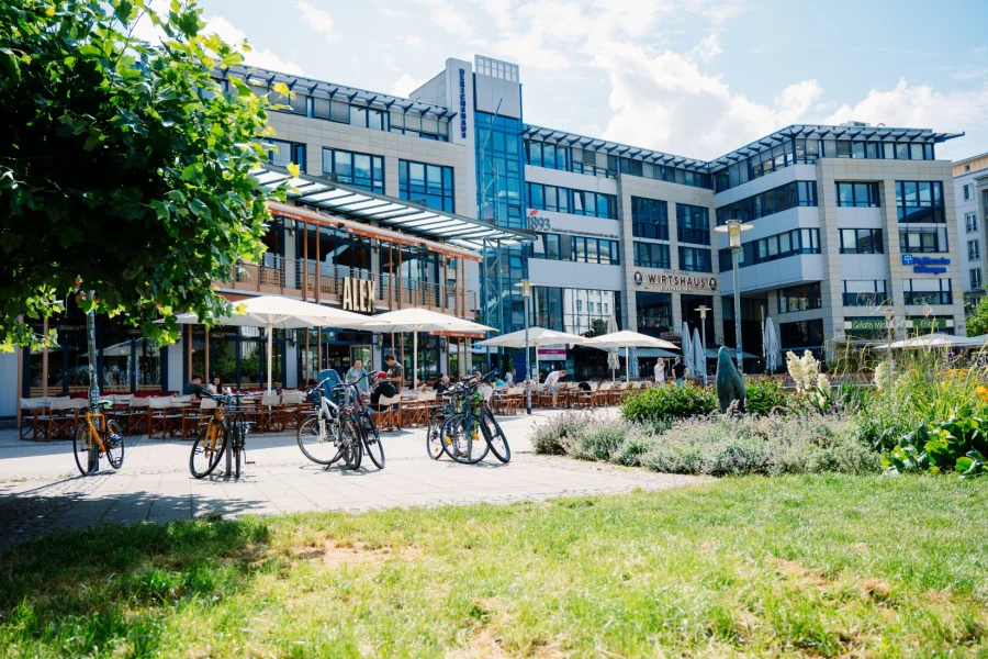 Terrasse des ALEX vor dem Ulrichhaus mit wei&szlig;en Sonnenschirmen, davor abgestellte Fahrr&auml;der und ein Staudenbeet mit B&auml;renskulptur