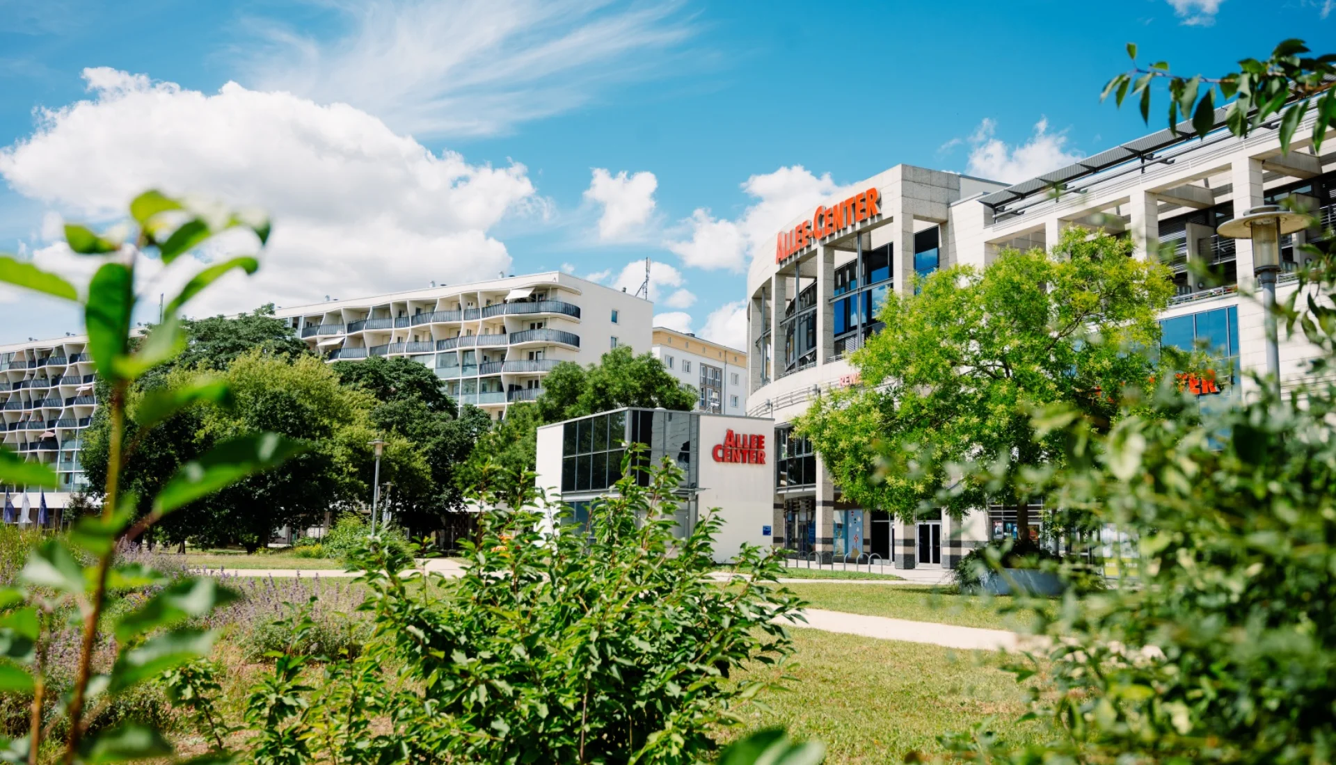 Allee-Center Magdeburg mit rotem Schriftzug &uuml;ber dem runden Eingangsbau, davor eine Wiese mit jungen Str&auml;uchern