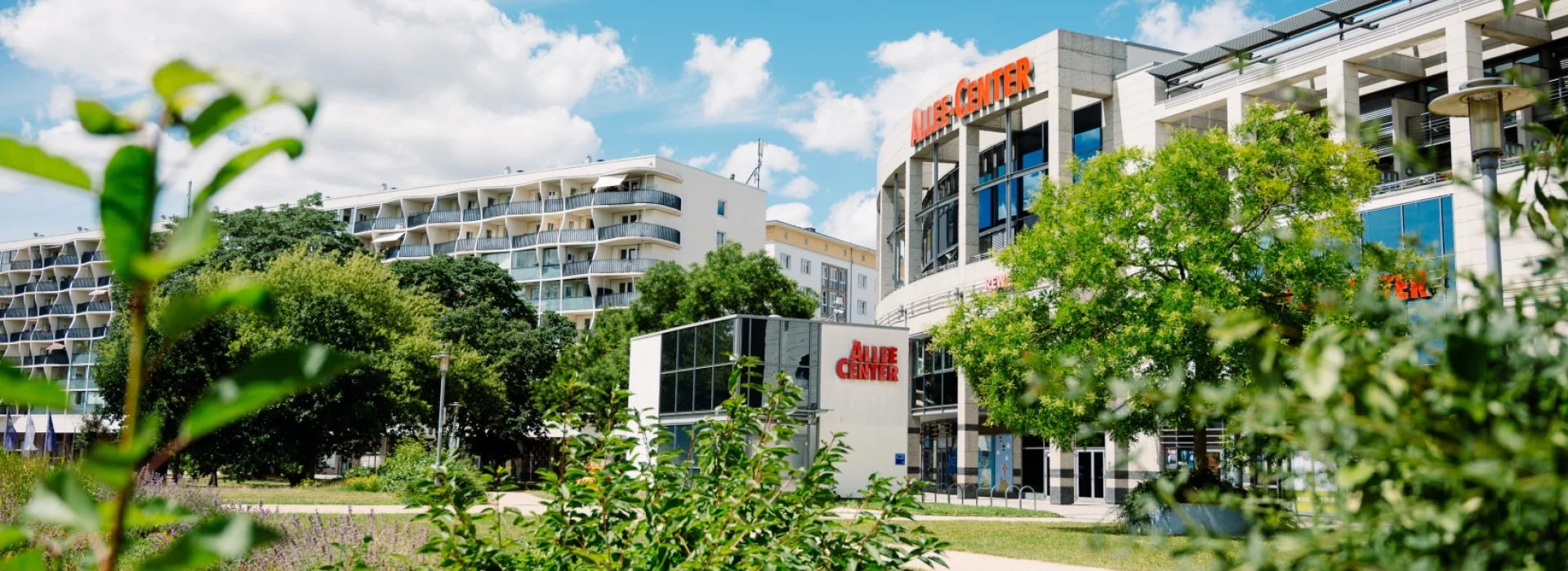 Allee-Center Magdeburg mit rotem Schriftzug über dem runden Eingangsbau, davor eine Wiese mit jungen Sträuchern