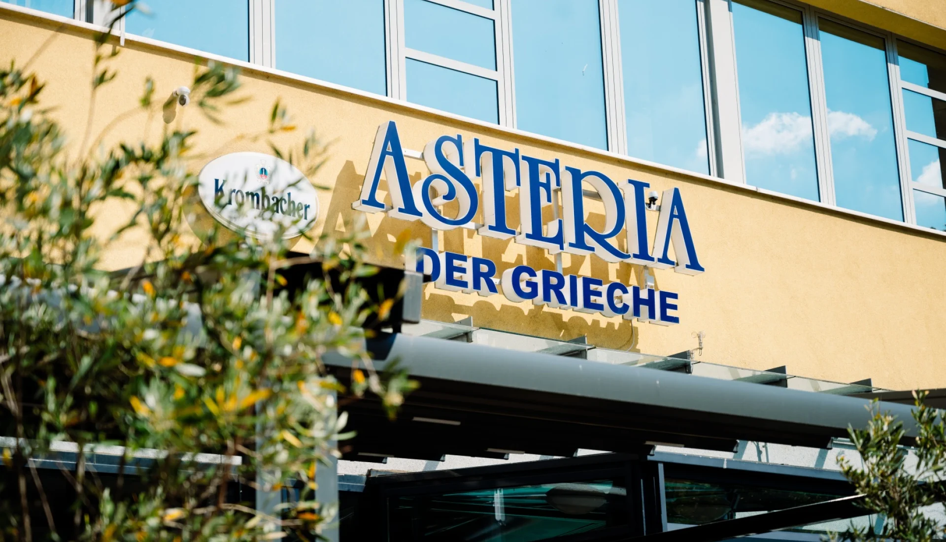 Blauer Schriftzug &bdquo;Asteria &ndash; Der Grieche&ldquo; an der gelben Fassade &uuml;ber dem Eingang, links davor die Zweige eines Olivenbaums