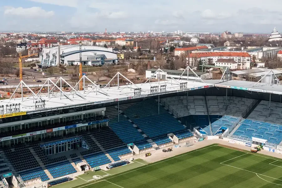 Luftaufnahme der Avnet Arena: leere blaue Tribuenen und gruener Rasen unter dem weissen Stahltraeger-Dach, dahinter das Stadtpanorama mit Jahrtausendturm