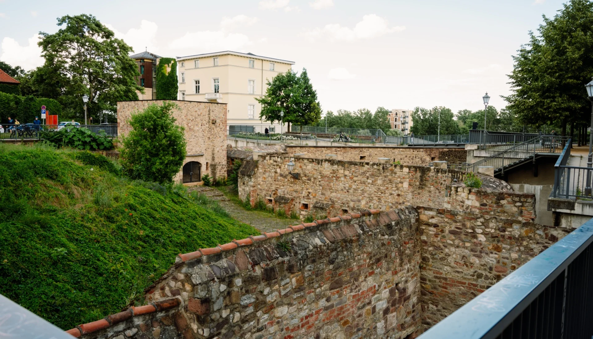 Festungsmauern der Bastion Cleve mit begruentem Wall und Wehrturm, dahinter eine cremefarbene Villa und Baeume am Fuerstenwall