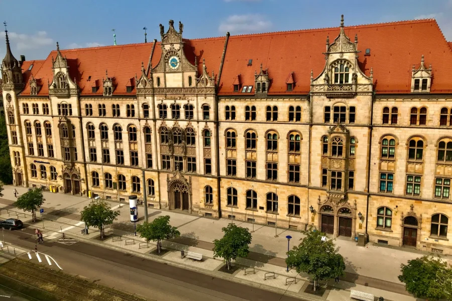 Die Alte Hauptpost am Breiten Weg: neogotischer Sandsteinbau mit rotem Ziegeldach, Uhrengiebel und Ziertuermchen, davor Baumreihe und Strassenbahntrasse