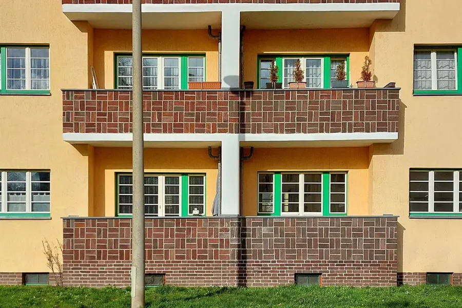 Fassade der Beimssiedlung: gelber Putz mit Backstein-Bruestungen, gruen gerahmte Sprossenfenster, orangefarbene Tueren und Balkone mit kleinen Baeumchen