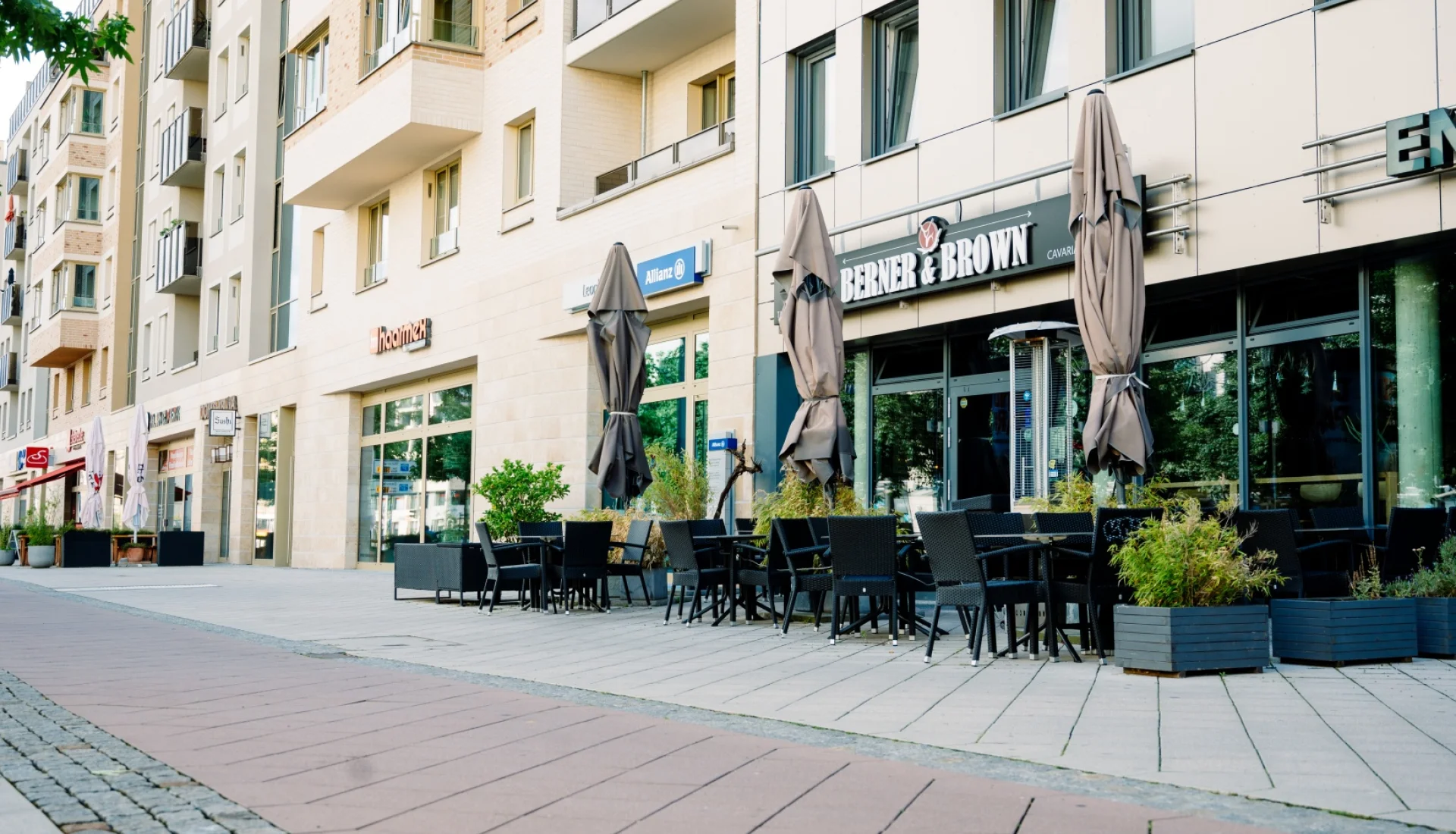 Stra&szlig;enterrasse des Berner & Brown mit dunklen Korbst&uuml;hlen, Pflanzk&uuml;beln und geschlossenen Sonnenschirmen vor dem Leuchtschriftzug