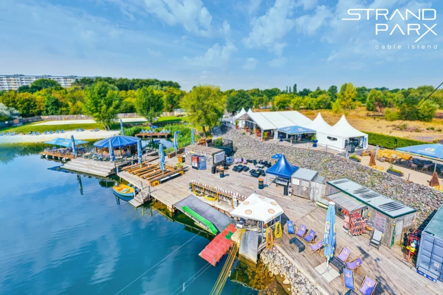 Luftaufnahme des STRANDPARX Cable Island am Neustaedter See: Holzstege mit Liegestuehlen und Sonnenschirmen, weisse Pagodenzelte und Sandstrand am tuerkisblauen Wasser