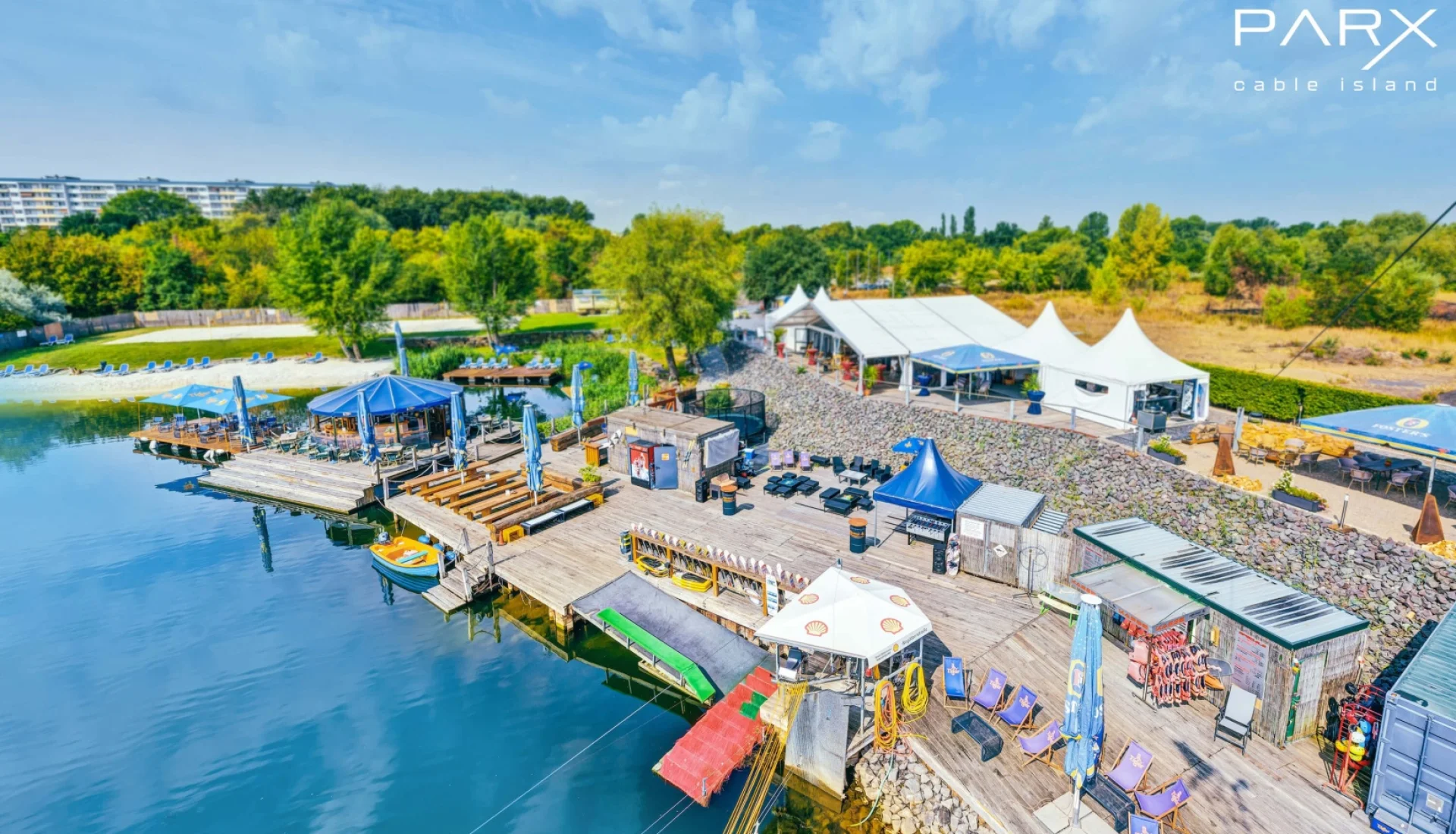 Cable Island aus der Luft: Holzstege mit Bars, Schirmen und Liegest&uuml;hlen am Wasserskisee, dahinter Sandstrand, wei&szlig;e Zelte und die Startanlage der Seilbahn