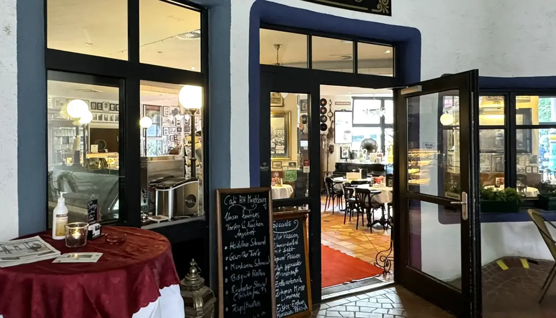 Blick in das Caf&eacute; Alt Magdeburg im Hundertwasserhaus: ge&ouml;ffnete Glast&uuml;r zwischen blau eingefassten Fenstern, drinnen Bistrotische auf Terrakottafliesen, links ein Stehtisch mit roter Decke