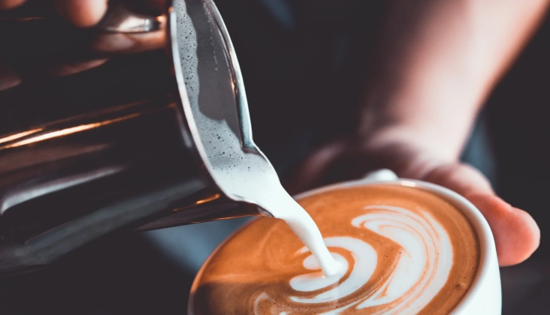 Barista gie&szlig;t Milchschaum aus einer Edelstahlkanne in eine Tasse Cappuccino mit Latte-Art-Muster, Nahaufnahme in warmem Licht