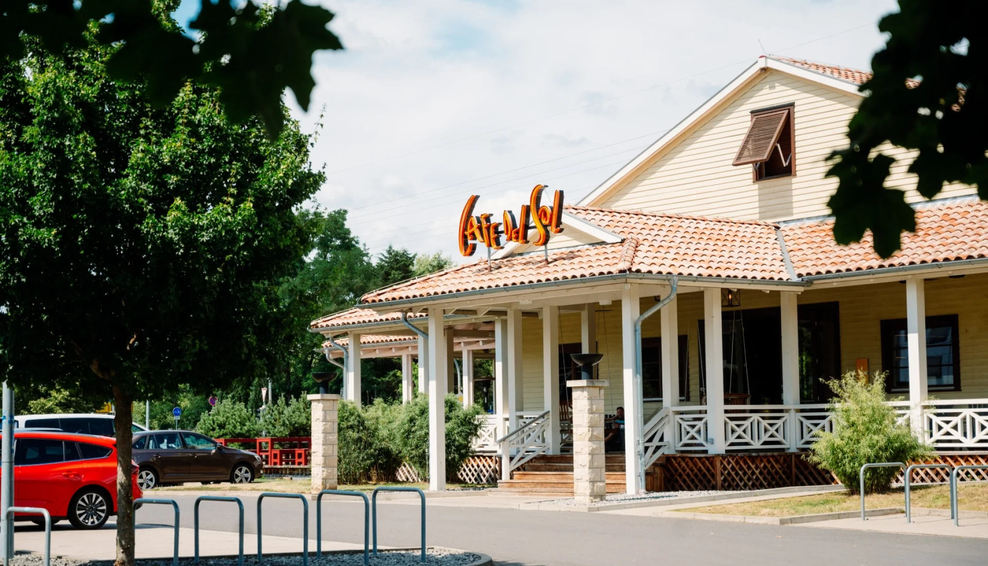 Cafe Del Sol Magdeburg von aussen: gelbes Holzhaus im Landhausstil mit rotem Ziegeldach, umlaufender Veranda mit weissem Gelaender und orangefarbenem Neon-Schriftzug auf dem Dach