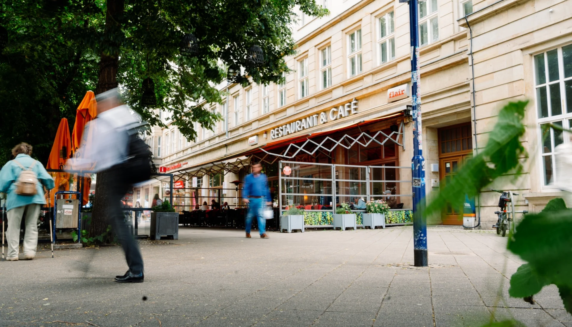 Das Flair am Boulevard: verglaste Terrasse unter der Markise, davor Passanten im Schatten der Stra&szlig;enb&auml;ume