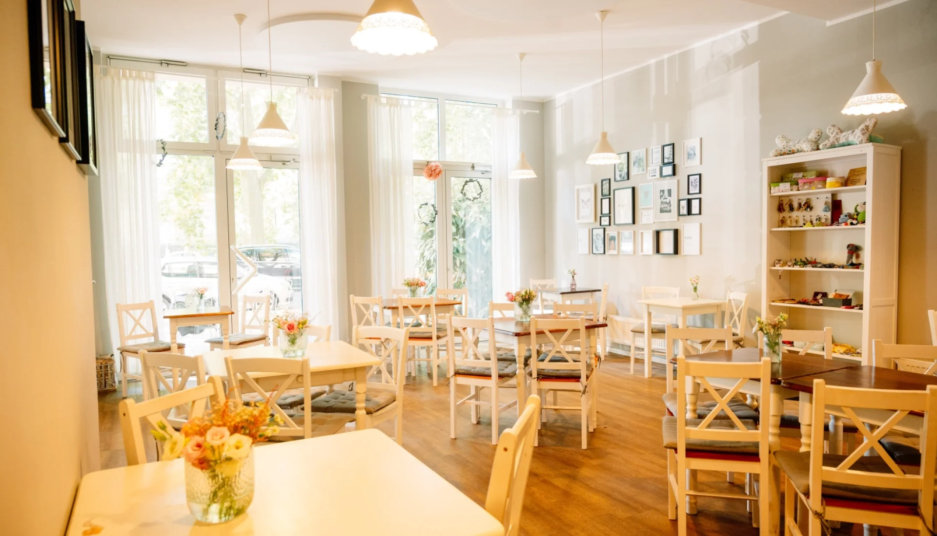 Gastraum des Cafe Herzstueck im Landhausstil: weisse Holztische und Sprossenstuehle mit Blumenstraeussen, Bilderwand und Regal mit Kindersachen, grosse Fenster mit weissen Vorhaengen
