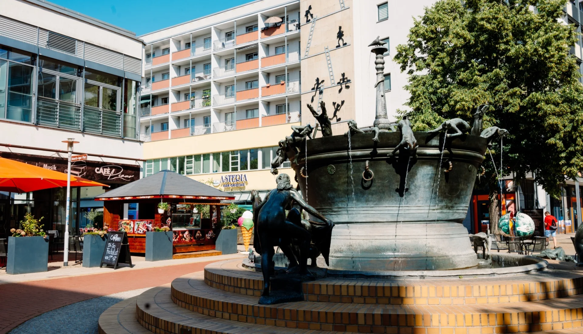 Faunenbrunnen mit seinen Bronzefiguren auf gestuftem Sockel, dahinter das Caf&eacute; Piano mit Terrasse und Sonnenschirmen
