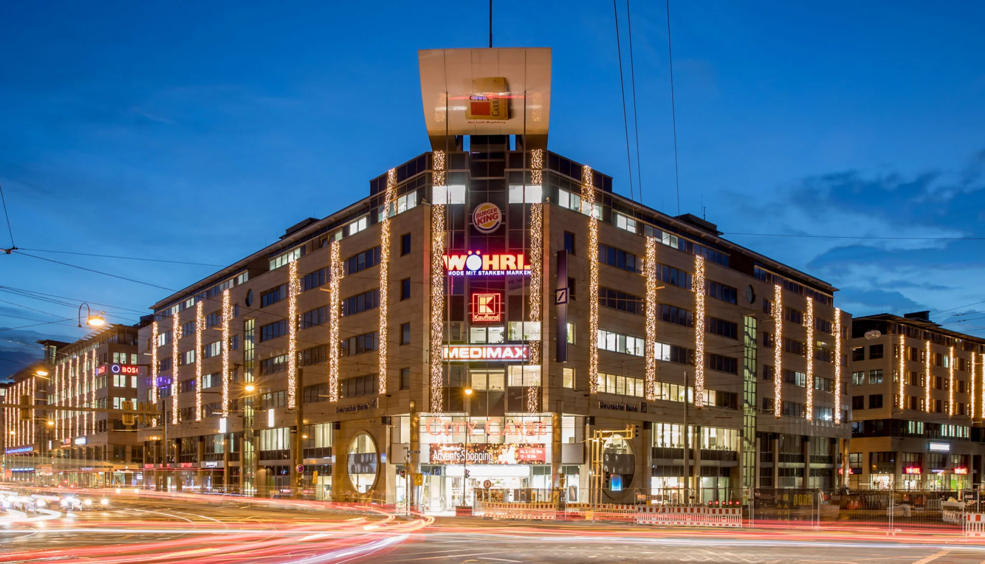 City Carr&eacute; zur blauen Stunde: Lichterketten an der Fassade, leuchtende Werbeschriften und Lichtspuren des Verkehrs davor