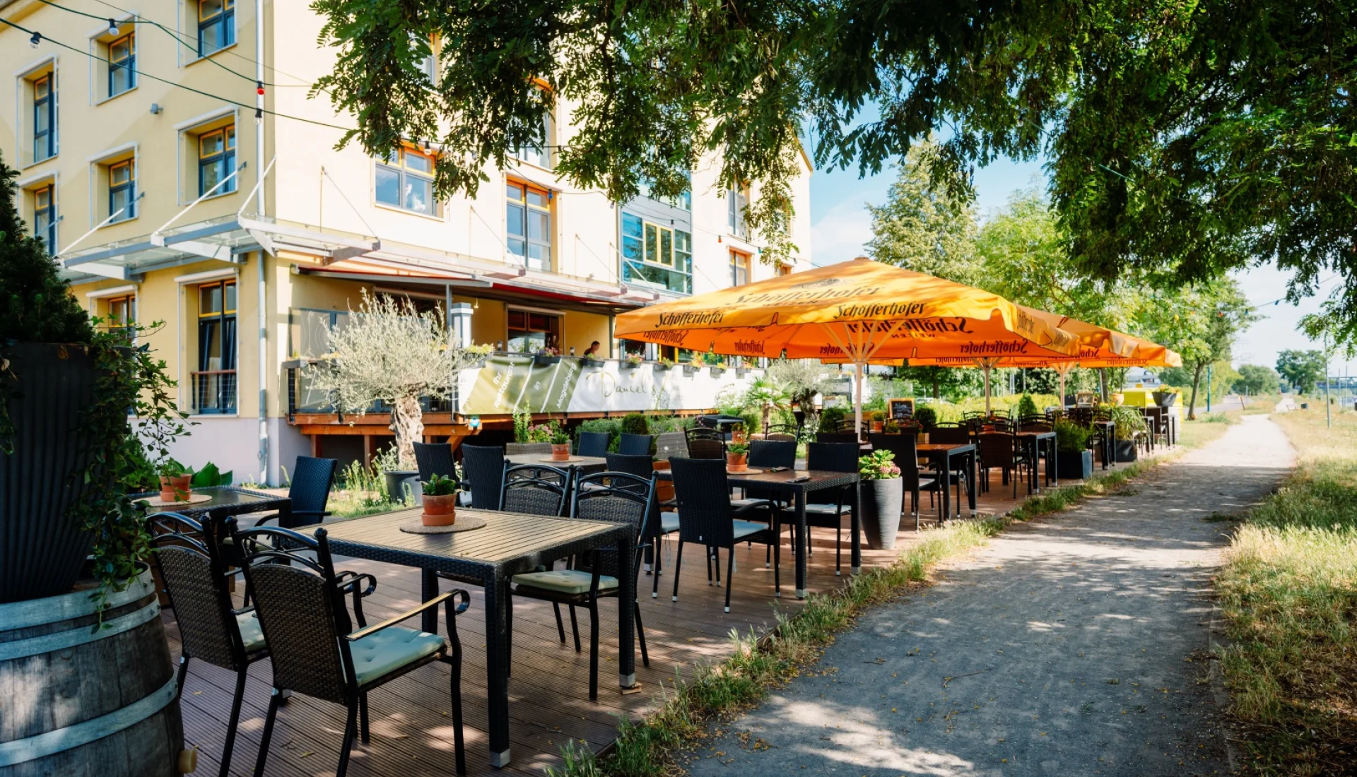 Terrasse des Daniel's am Elbradweg: schwarze Rattanst&uuml;hle und Holztische unter orangefarbenen Sonnenschirmen, dahinter das gelbe Elbwerk-Geb&auml;ude