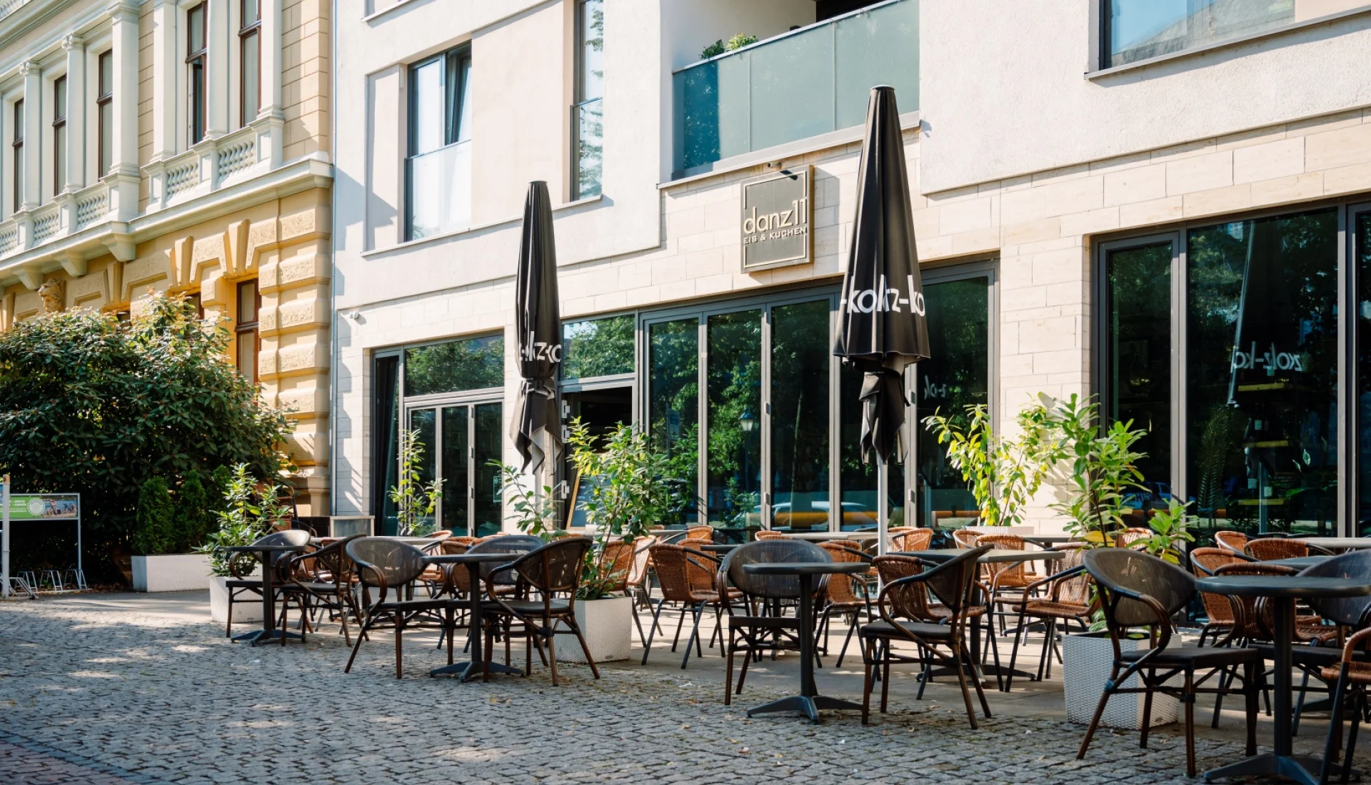 Terrasse des danz11 mit dunklen Korbst&uuml;hlen und zusammengebundenen Sonnenschirmen vor der Glasfront, links schlie&szlig;t eine Gr&uuml;nderzeitfassade an