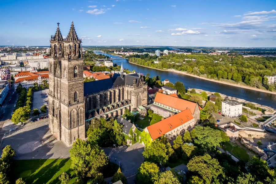 Luftaufnahme des Magdeburger Doms mit Kreuzgang und rotem Klosterdach, dahinter die Elbe mit Strombruecke und der gruene Rotehornpark