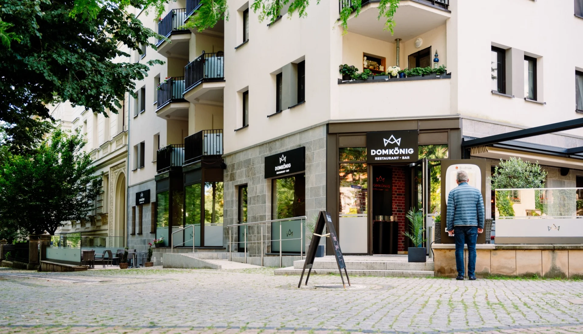 Eckgeb&auml;ude mit dem Domk&ouml;nig im Erdgeschoss: dunkle Ladenfront mit Logo &uuml;ber Eingang und Terrasse, davor ein gepflasterter Platz mit Aufsteller