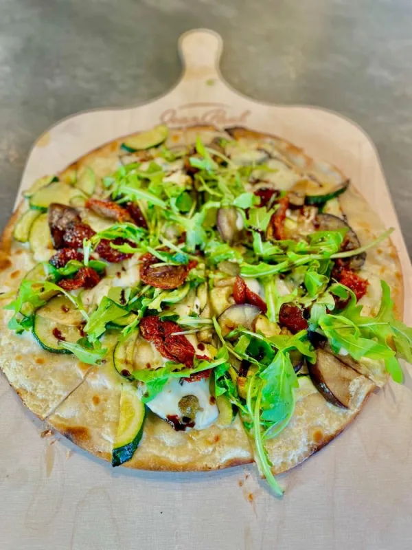 Dünner Flammkuchen mit Zucchini, Aubergine, getrockneten Tomaten, Mozzarella und Rucola auf einem hellen Holzschieber