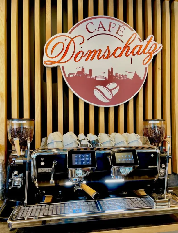 Siebträgermaschine mit vorgewärmten Tassen vor einer Holzlattenwand, darüber das Logo des Café Domschatz