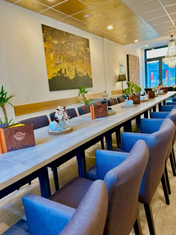 Lange Tafel mit blauen Polsterstühlen im Gastraum des Café Domschatz, an der Wand ein goldfarbenes Bild mit dem Café-Schriftzug