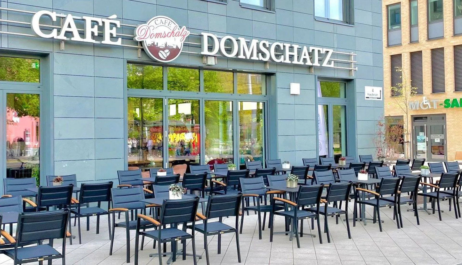 Terrasse des Caf&eacute; Domschatz mit Reihen dunkelblauer St&uuml;hle und Tische vor der grauen Fassade, &uuml;ber der Glasfront der Schriftzug des Caf&eacute;s