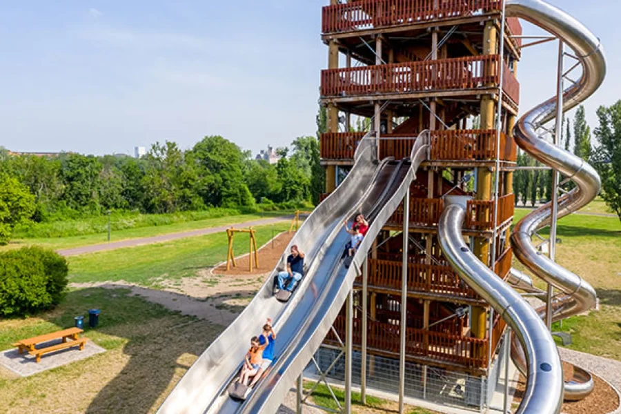 Kinder und Erwachsene rutschen auf den breiten Edelstahlrutschen des h&ouml;lzernen Rutschenturms im Elbauenpark abw&auml;rts