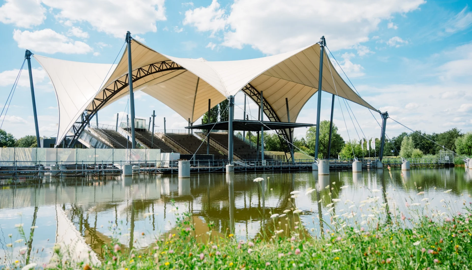 Seeb&uuml;hne im Elbauenpark mit ihren wei&szlig;en Zeltd&auml;chern und der leeren Trib&uuml;ne, gespiegelt im See, davor eine Blumenwiese