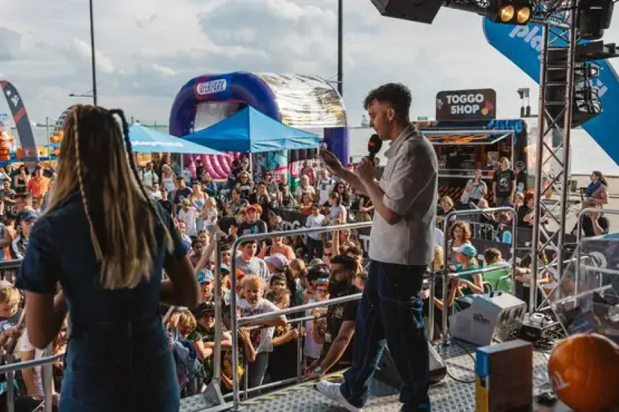 Buehne der TOGGO Tour am Elbufer: zwei Moderierende vor einer dicht gefuellten Zuschauerflaeche mit Huepfburgen und TOGGO-Shop