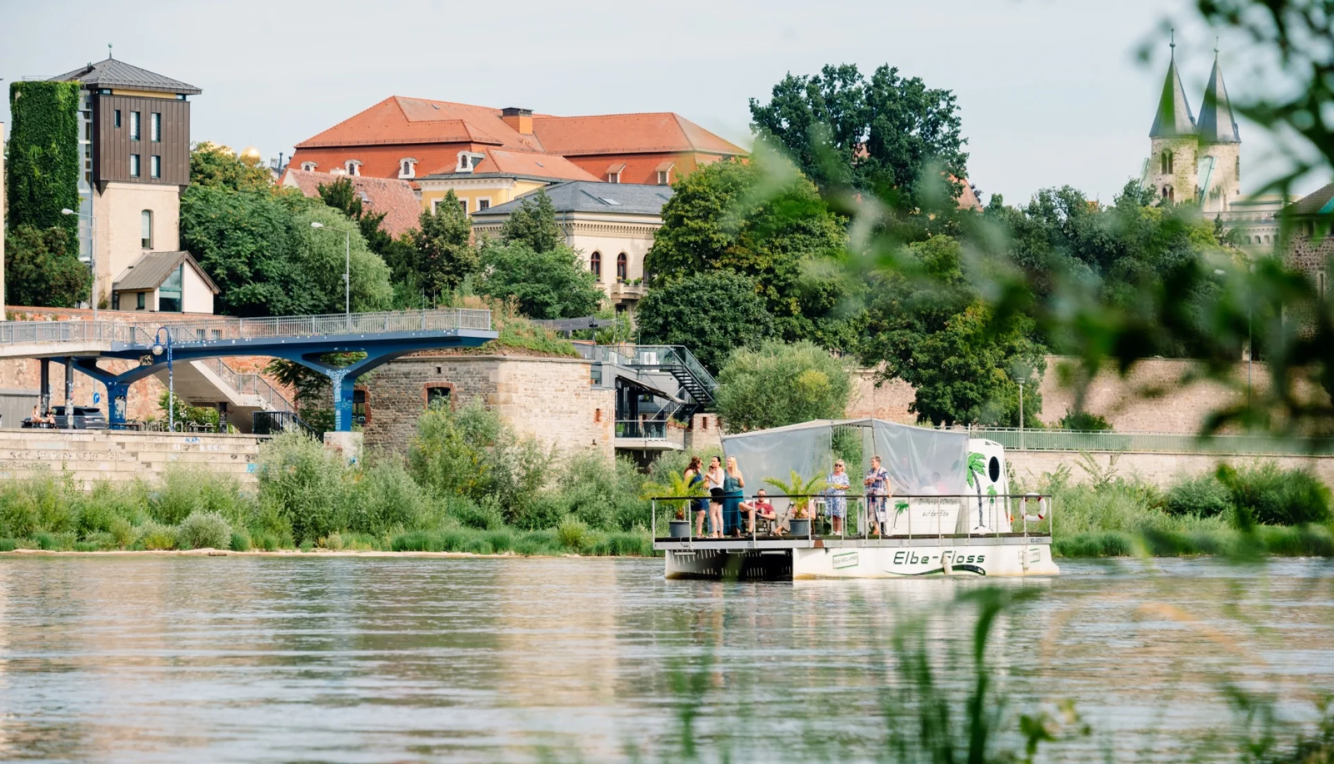 Elbe-Flo&szlig; mit G&auml;sten und Palmen an Deck auf der Elbe, dahinter eine blaue Fu&szlig;g&auml;ngerbr&uuml;cke und die T&uuml;rme des Klosters Unser Lieben Frauen