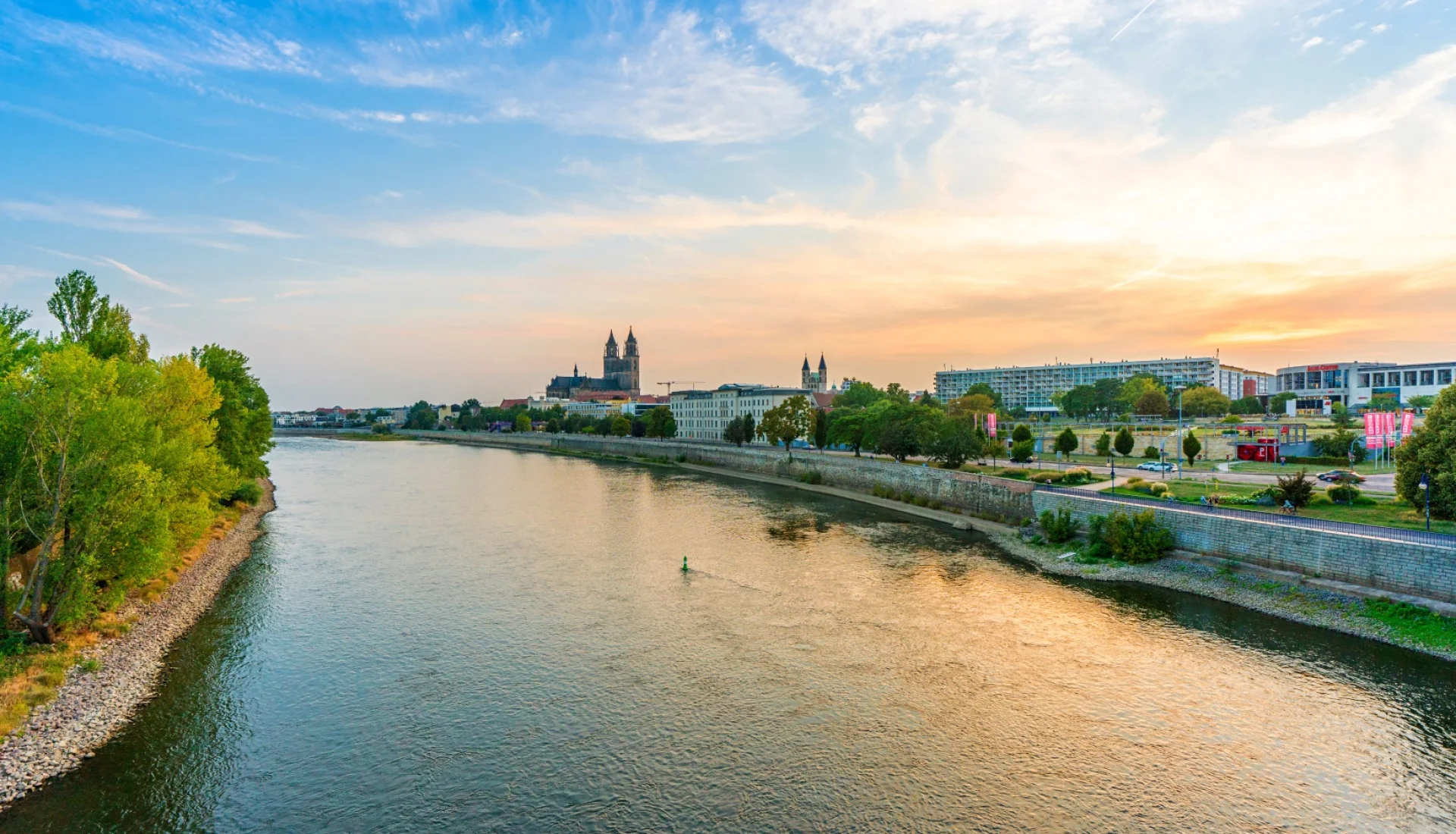 Abendlicher Blick ueber die Elbe auf Magdeburg: links bewaldetes Ufer, in der Mitte die Domtuerme ueber der Uferpromenade im goldenen Licht