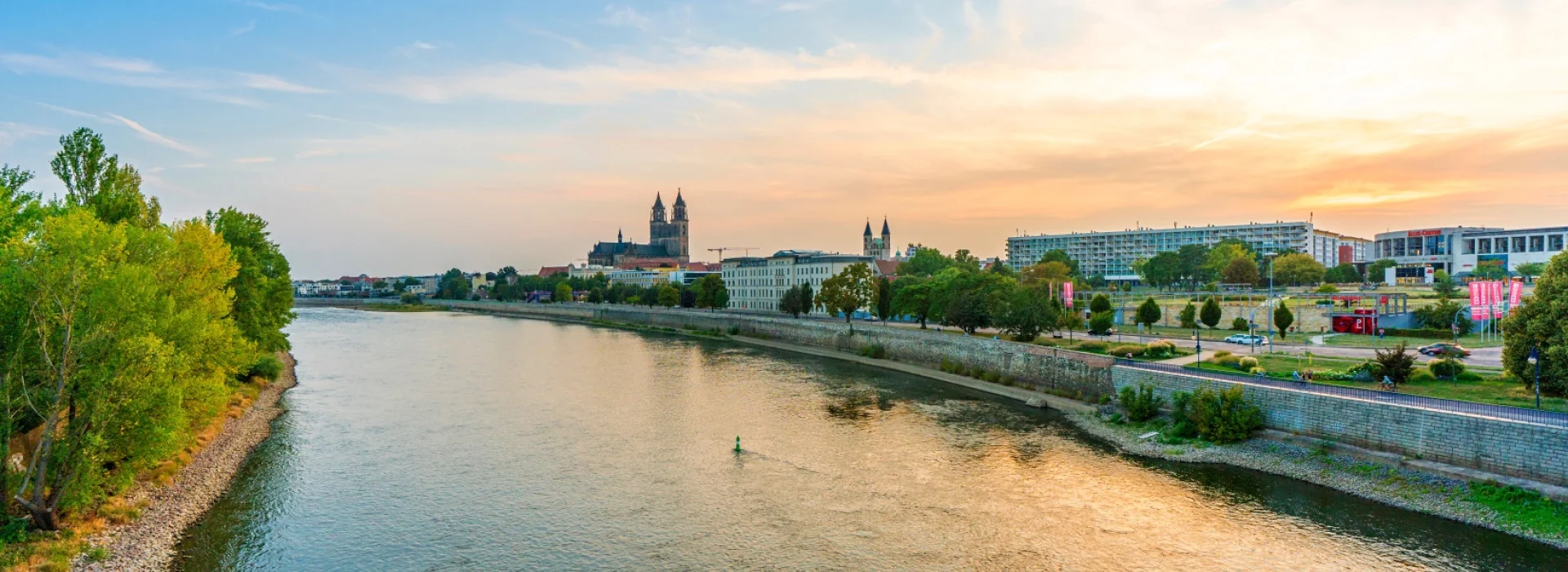 Abendlicher Blick ueber die Elbe auf Magdeburg: links bewaldetes Ufer, in der Mitte die Domtuerme ueber der Uferpromenade im goldenen Licht
