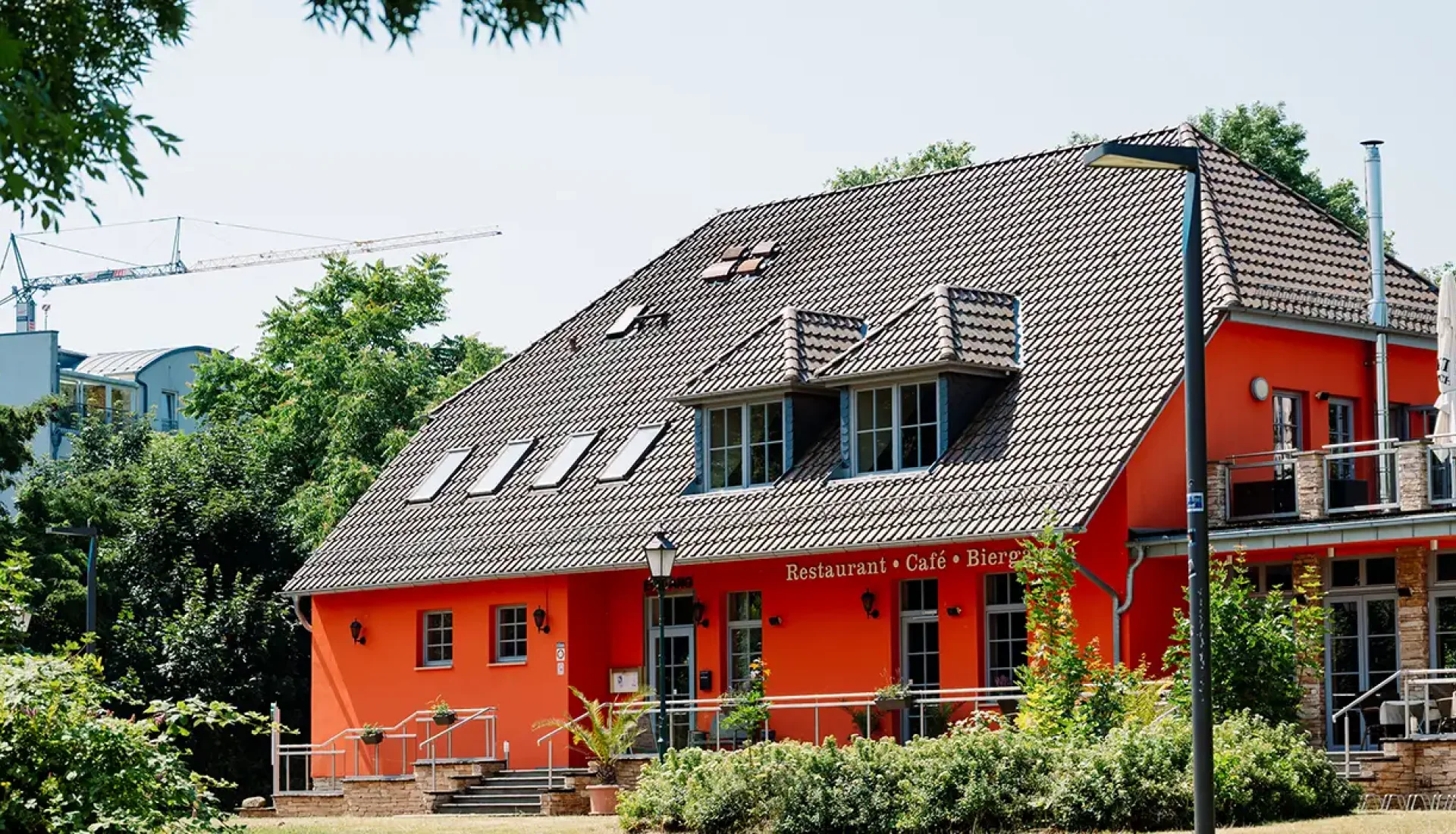 Rotes Elbelandhaus mit Dachgauben und Schornstein hinter Str&auml;uchern, rechts die erh&ouml;hte Terrasse mit Sonnenschirmen, links ein Baukran am Himmel
