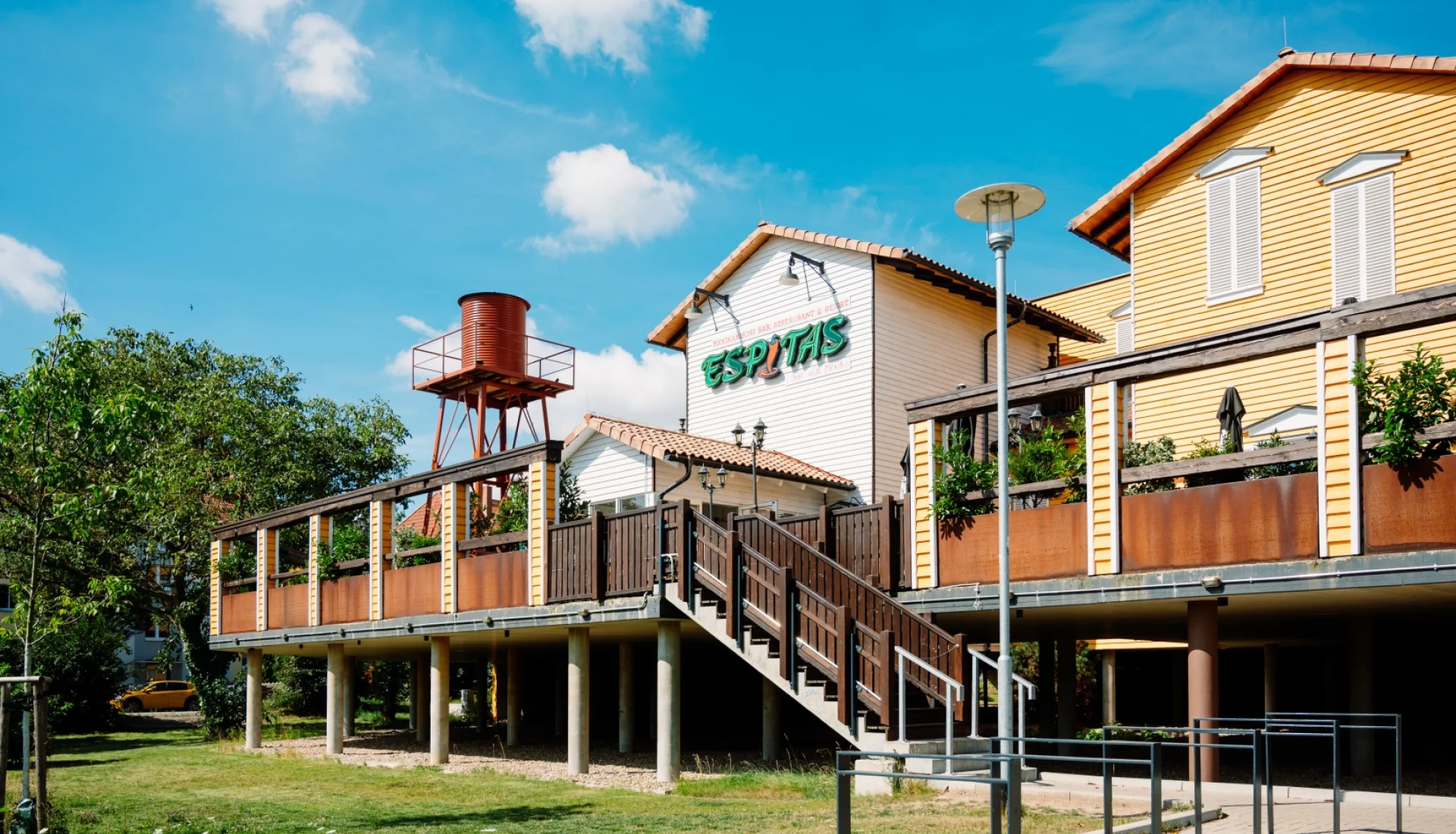 Aussenansicht des Espitas Magdeburg: erhoehte Holzterrasse auf Betonstelzen mit Treppenaufgang, dahinter gelbe Holzhaeuser und ein rostroter Wasserturm unter blauem Himmel