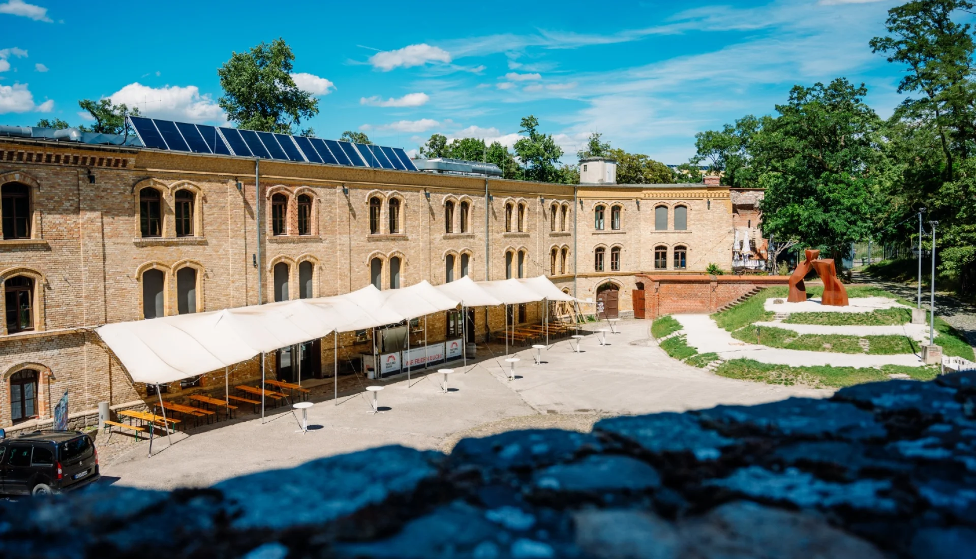 Innenhof der Festung Mark von der Mauer aus: langes Kasernengebaeude aus gelbem Backstein mit Solardach, davor weisses Sonnensegel ueber Biertischgarnituren und Stehtischen, rechts eine rostrote Skulptur auf der Wiese