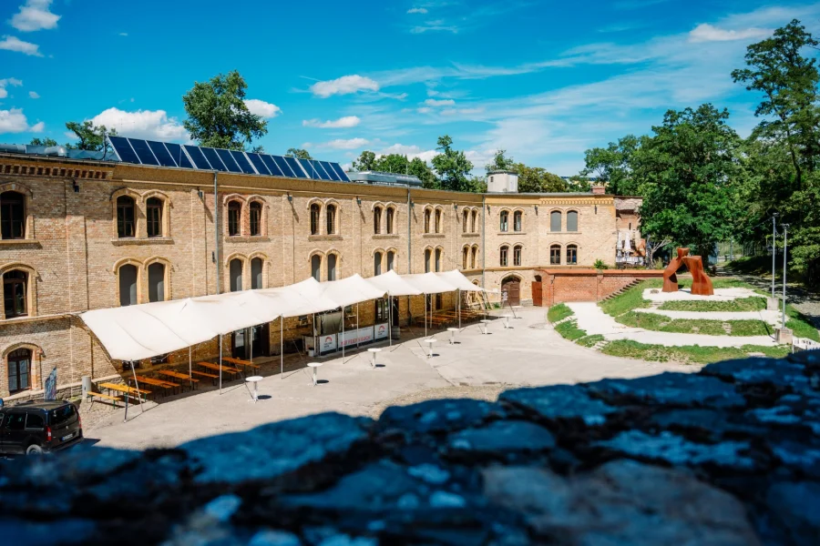 Innenhof der Festung Mark von der Mauer aus: langes Kasernengebaeude aus gelbem Backstein mit Solardach, davor weisses Sonnensegel ueber Biertischgarnituren und Stehtischen, rechts eine rostrote Skulptur auf der Wiese