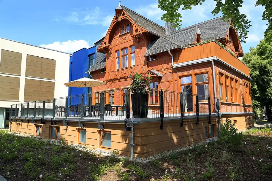 Saniertes Rayonhaus mit rot-orangefarbener Holzfassade, verziertem Giebel und vorgelagerter Terrasse, daneben moderne Neubauten
