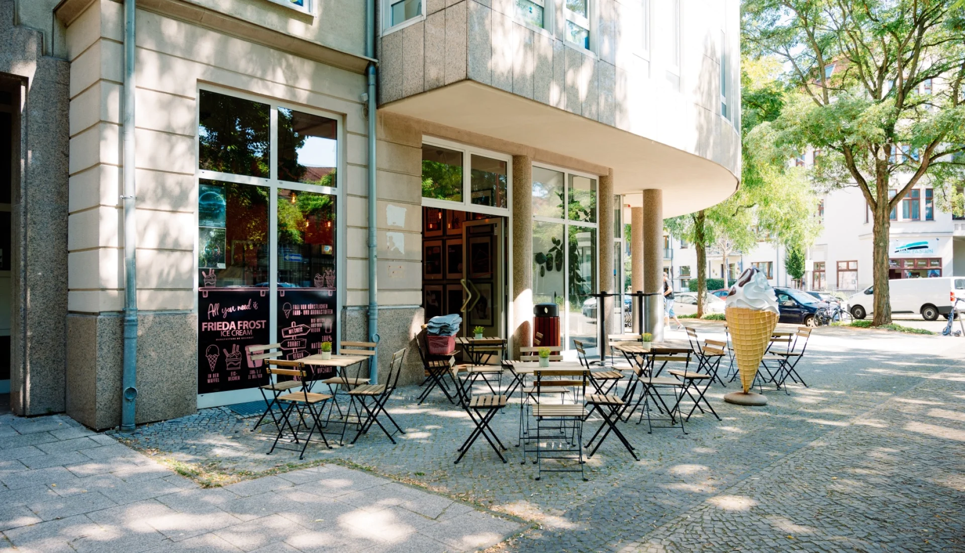 Aussenbereich von Frieda Frost an einer Gebaeudeecke: Klapptische und Stuehle auf dem Gehweg im Baumschatten, am Schaufenster die rosa Speisekarte, rechts eine grosse Eistuete als Aufsteller