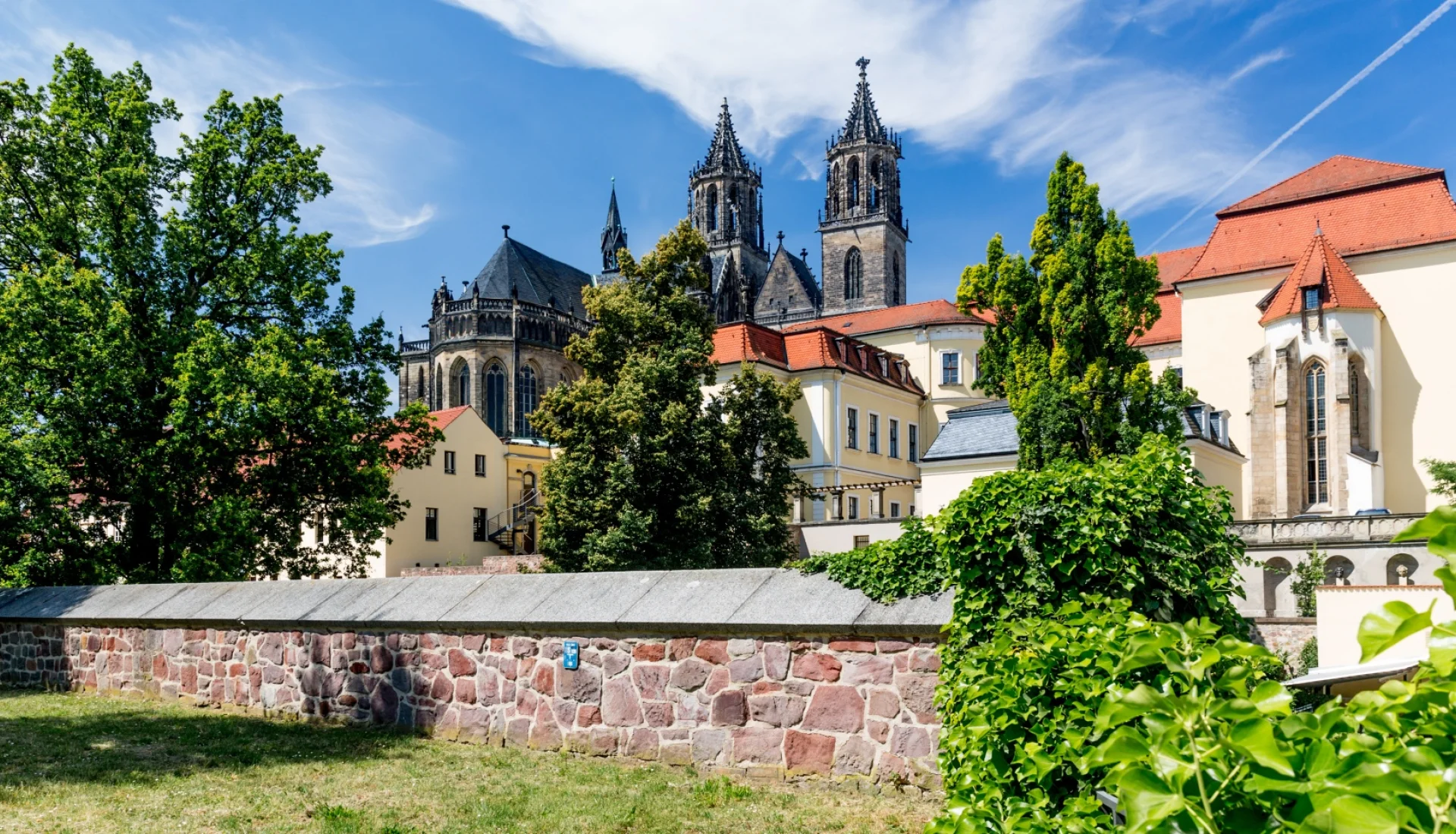Blick ueber die rote Bruchsteinmauer am Fuerstenwall auf Dom und Domklausur mit ihren roten Daechern unter blauem Sommerhimmel