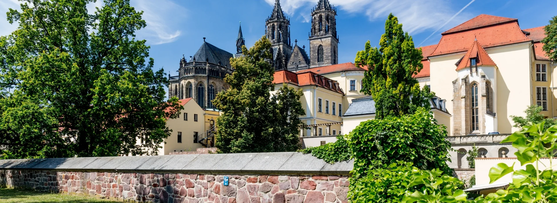 Blick ueber die rote Bruchsteinmauer am Fuerstenwall auf Dom und Domklausur mit ihren roten Daechern unter blauem Sommerhimmel