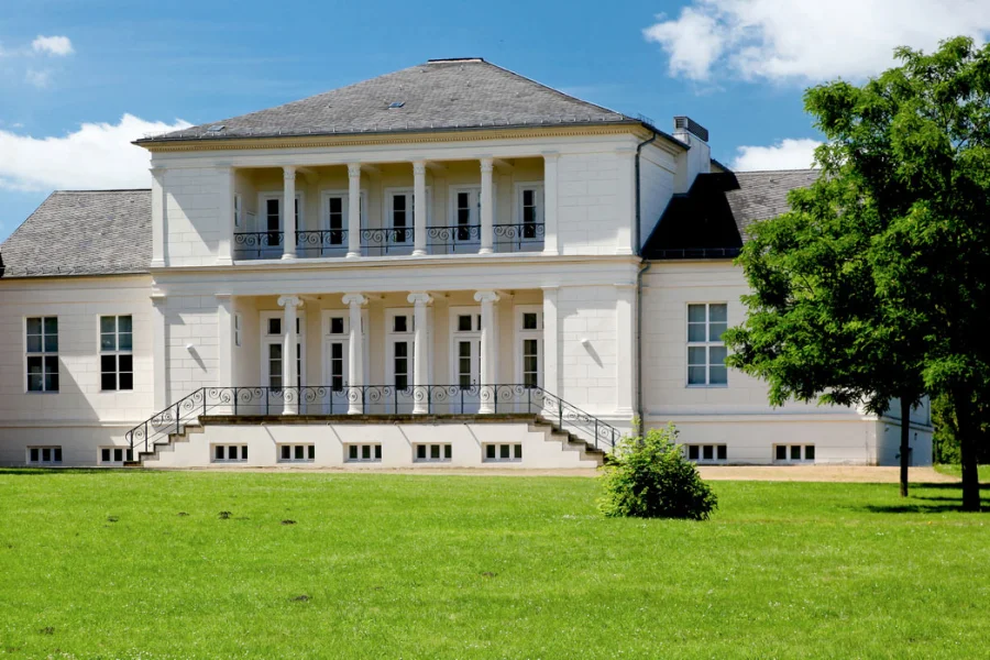 Das klassizistische Gesellschaftshaus im Klosterbergegarten: weisser Bau mit Saeulenportikus und Freitreppe hinter grosser Rasenflaeche, rechts ein Baum im Sommerlaub