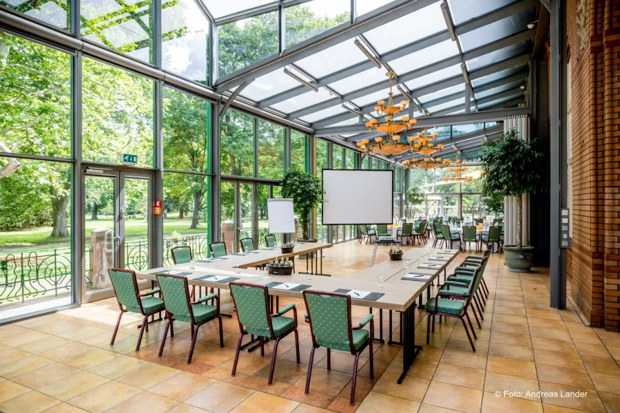Tagung im verglasten Wintergarten: lange Tafel mit gr&uuml;n gepolsterten St&uuml;hlen, Leinwand und Blick in den Park