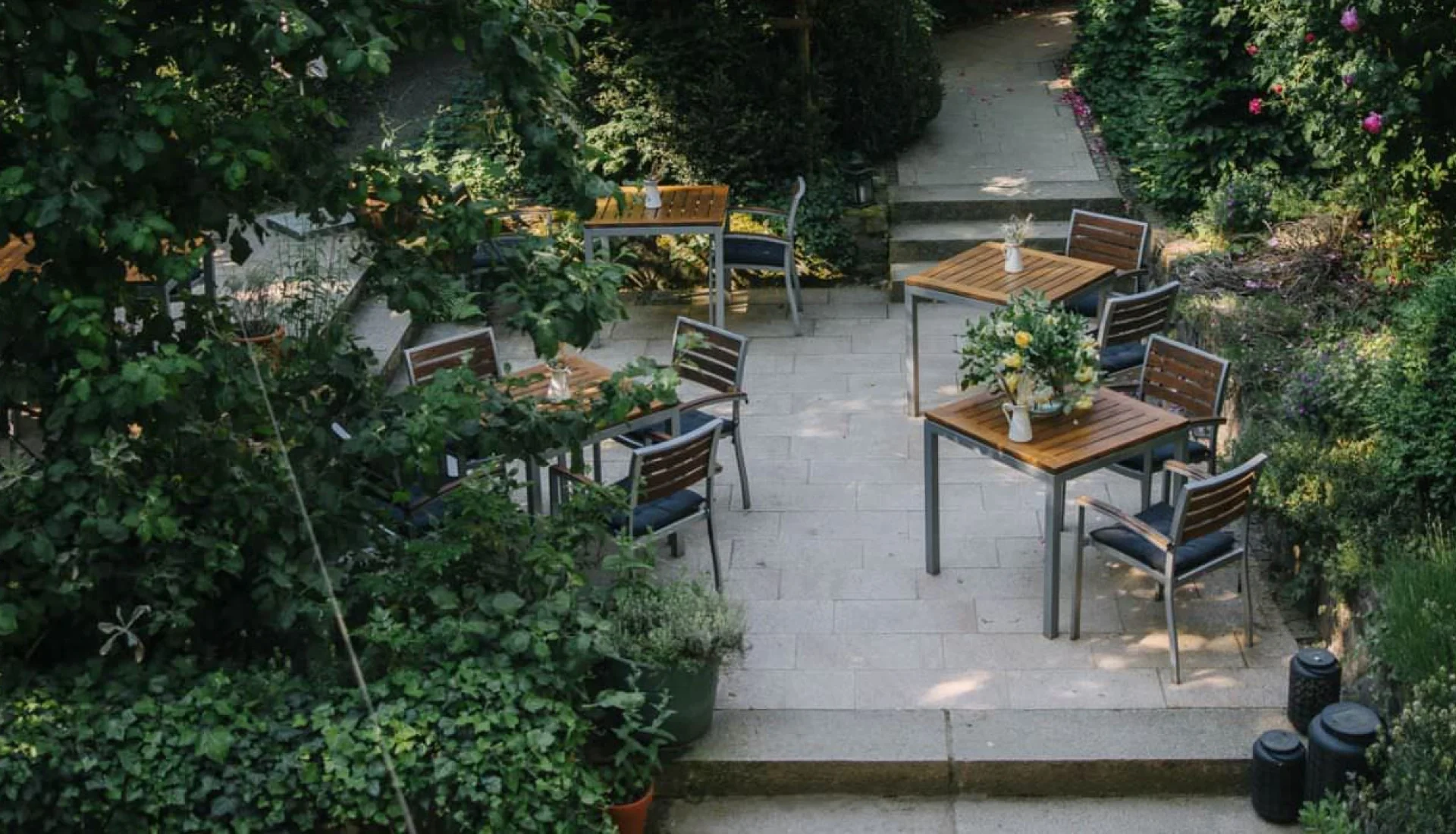 Terrassengarten des Hoflieferant von oben: Holztische mit Blumenstr&auml;u&szlig;en auf gestuften Steinplatten, eingefasst von Hecken, Rosen und gelben Dahlien